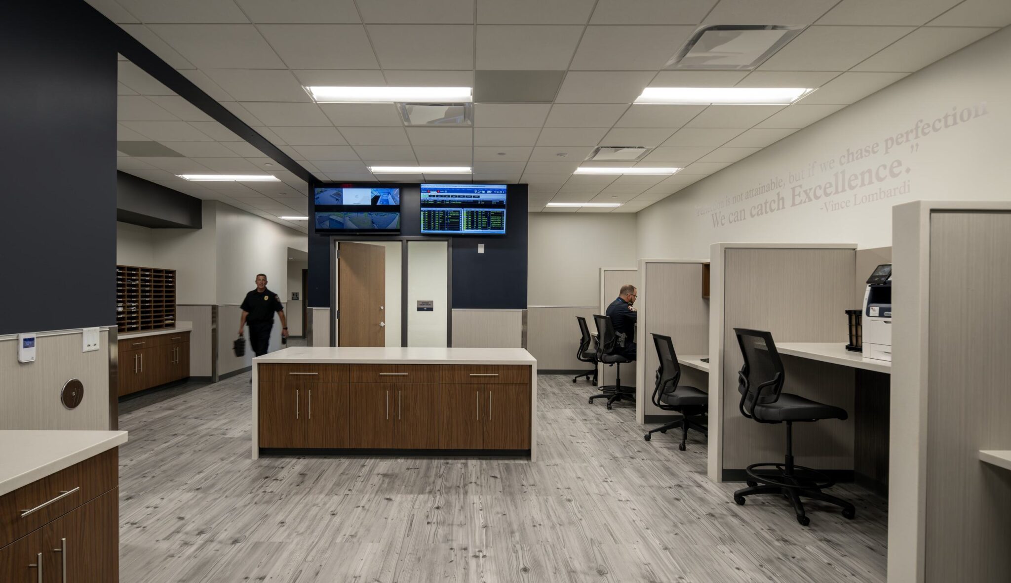 A modern office space with wooden cabinetry, workstations, and monitors, featuring two people engaged in activities within a bright environment.