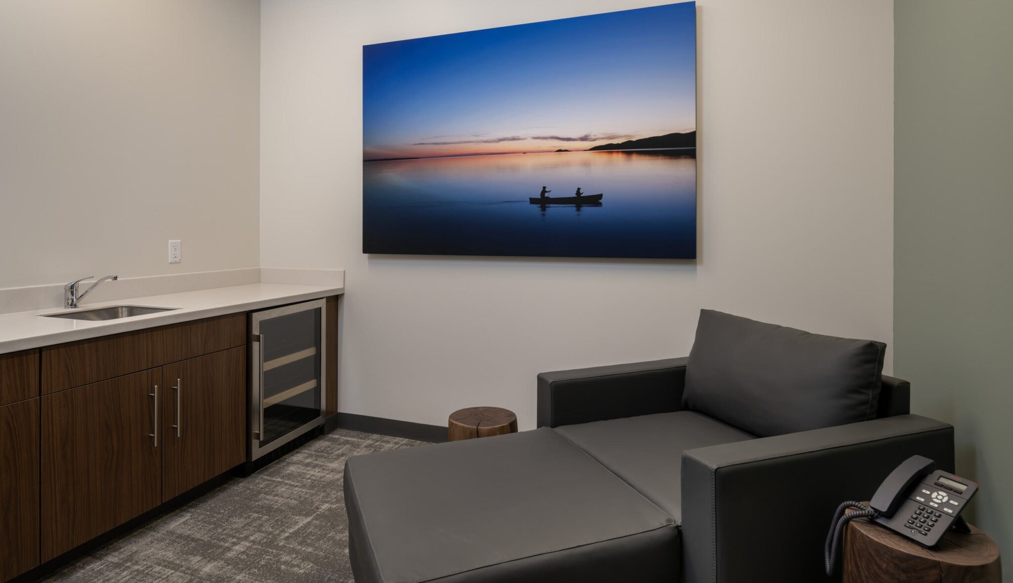 A serene room featuring a modern lounge chair, a kitchenette, and a tranquil landscape painting of a sunset over water.