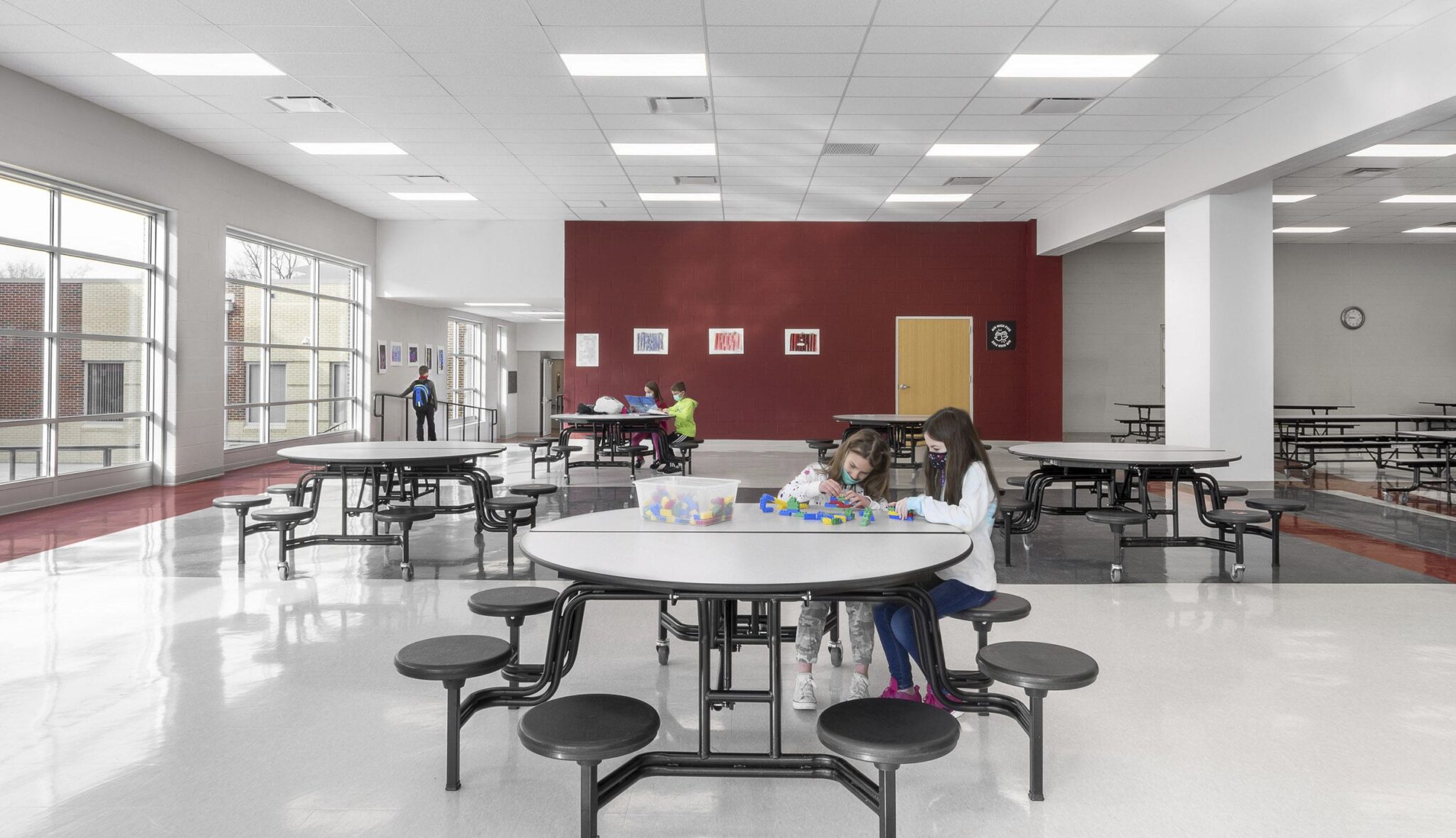 A spacious, modern cafeteria features empty tables and a few children engaged in activities, with large windows letting in natural light.