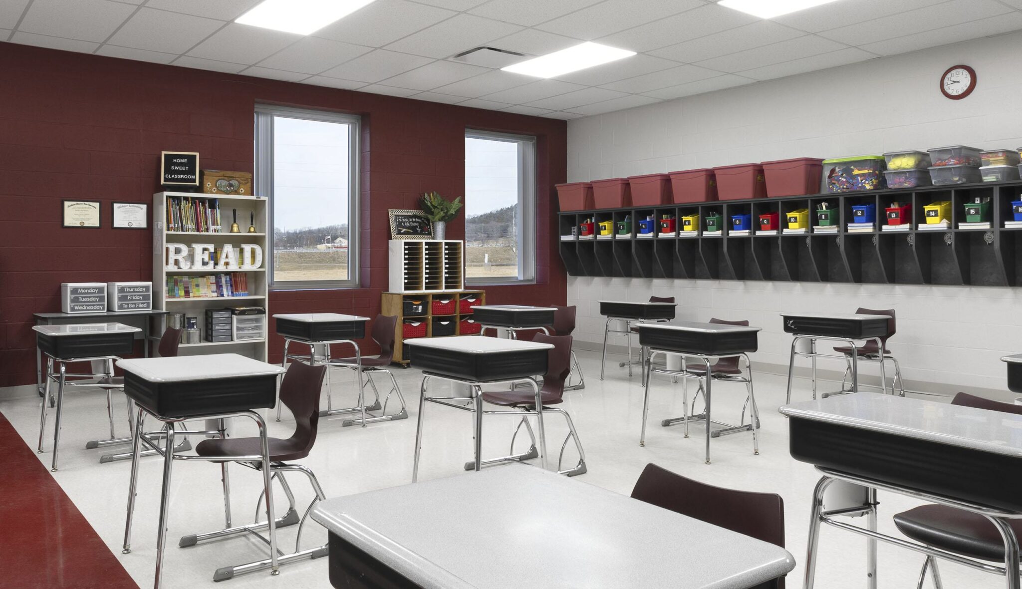 A modern, brightly lit classroom features rows of desks, a reading corner, and colorful storage bins along the wall. Large windows allow natural light.