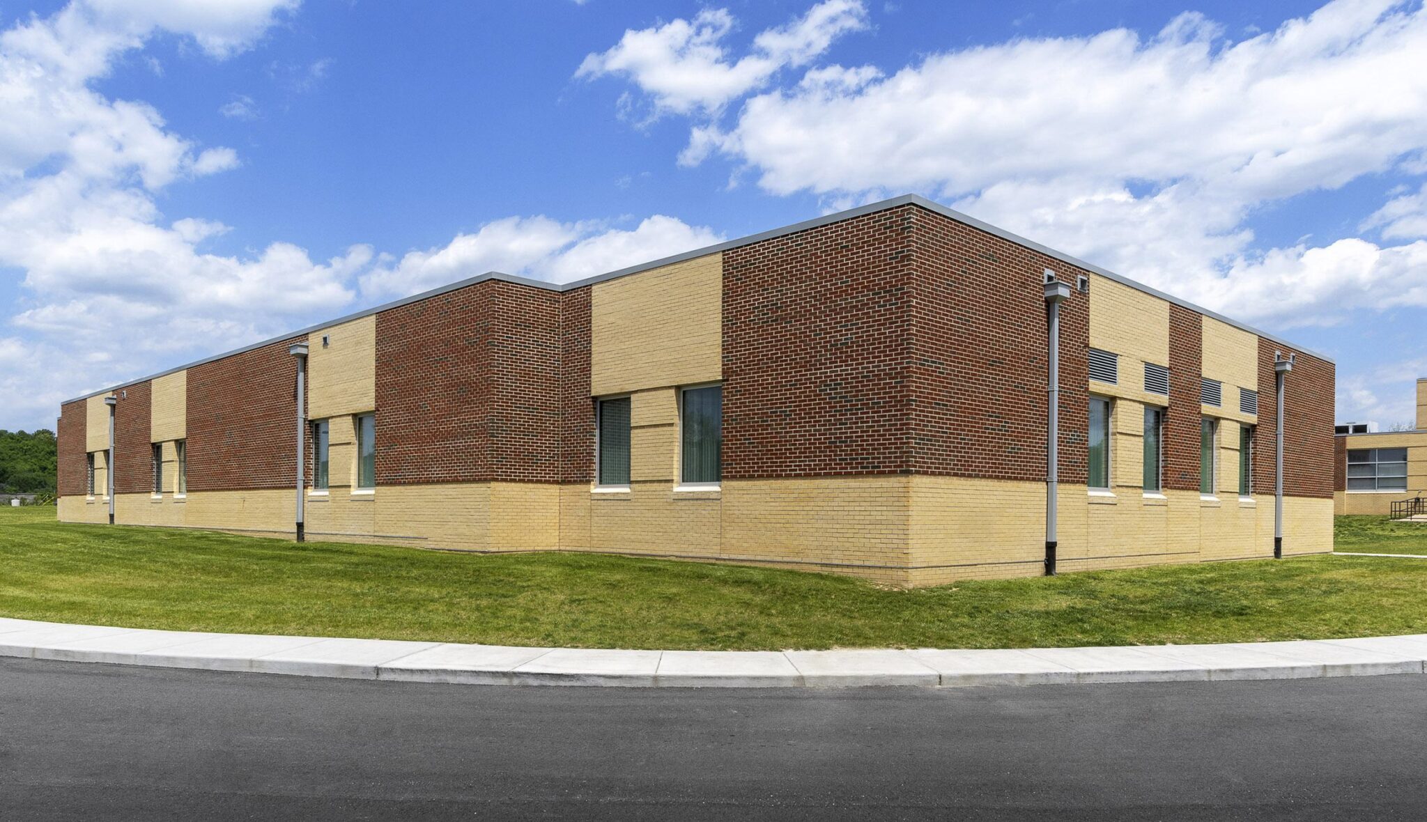 A modern, low-rise building features a mix of tan and brown brick, set against a blue sky with fluffy clouds, surrounded by green grass.