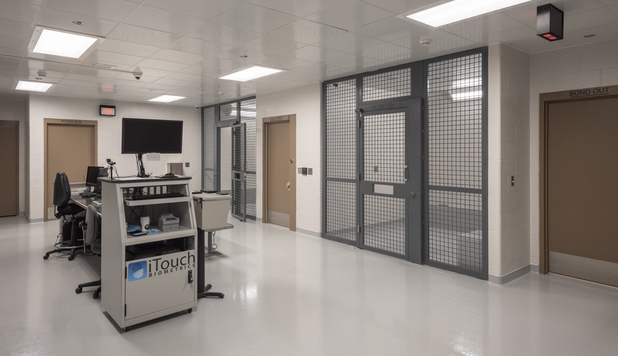 A modern, sterile hallway features secured doors, a computer station, and a monitoring area, with bright overhead lighting.