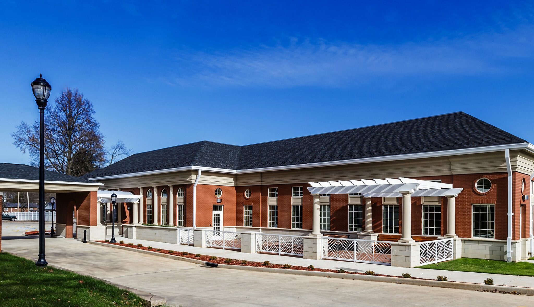 A modern brick building with large windows, a white pergola, and landscaped surroundings, set against a clear blue sky.