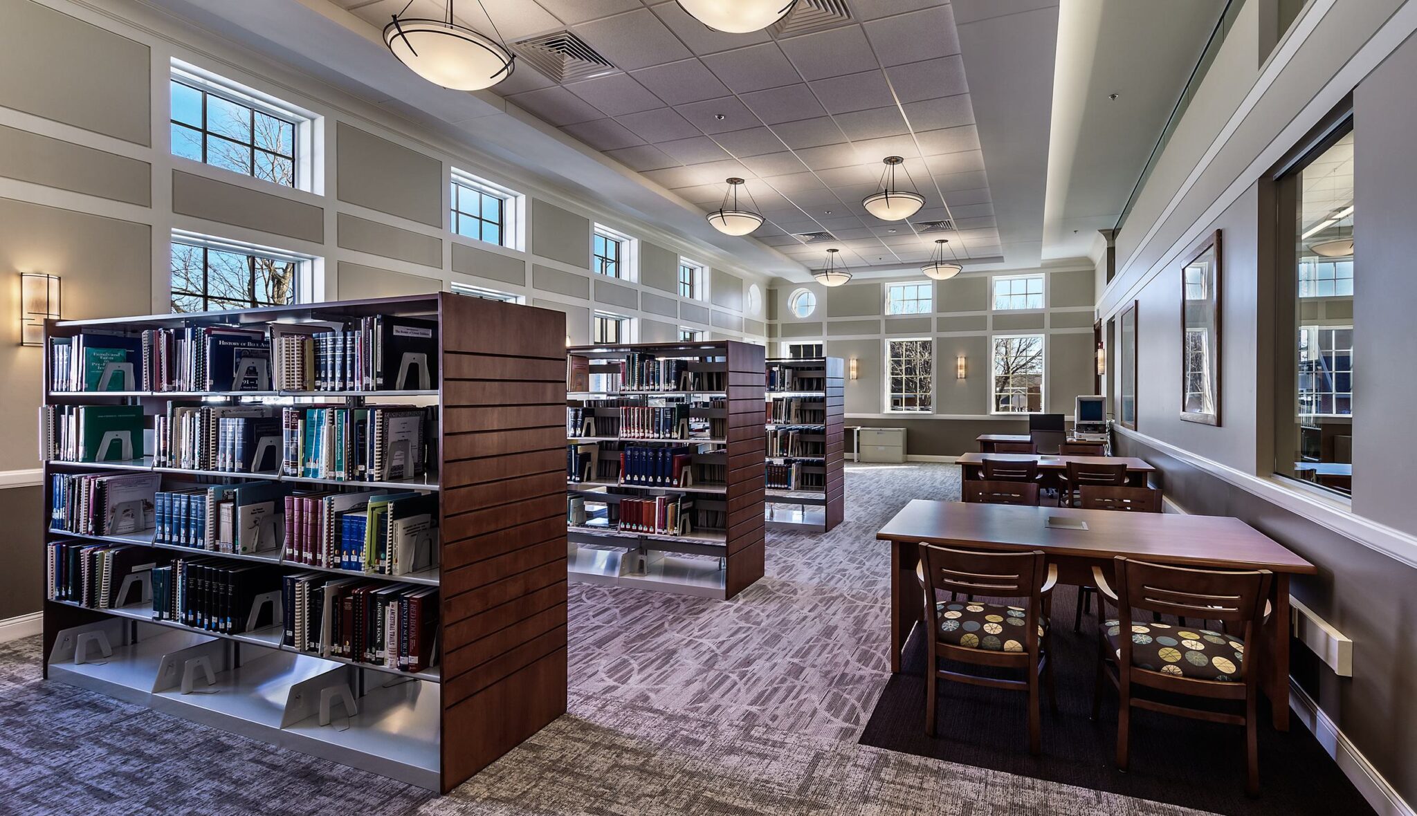 A bright and spacious library features wooden bookshelves, multiple tables for study, and large windows letting in natural light.