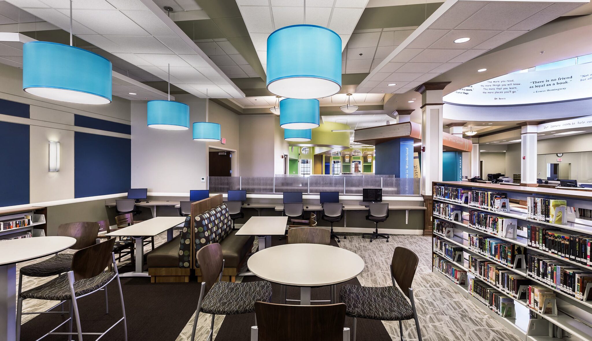 A modern library interior features bright pendant lighting, study tables, comfortable seating, and shelves filled with books.