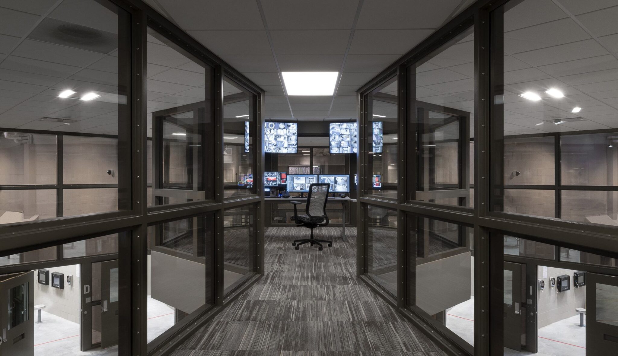 A modern control room features a central workspace surrounded by large glass windows, overlooking other areas equipped with monitors.