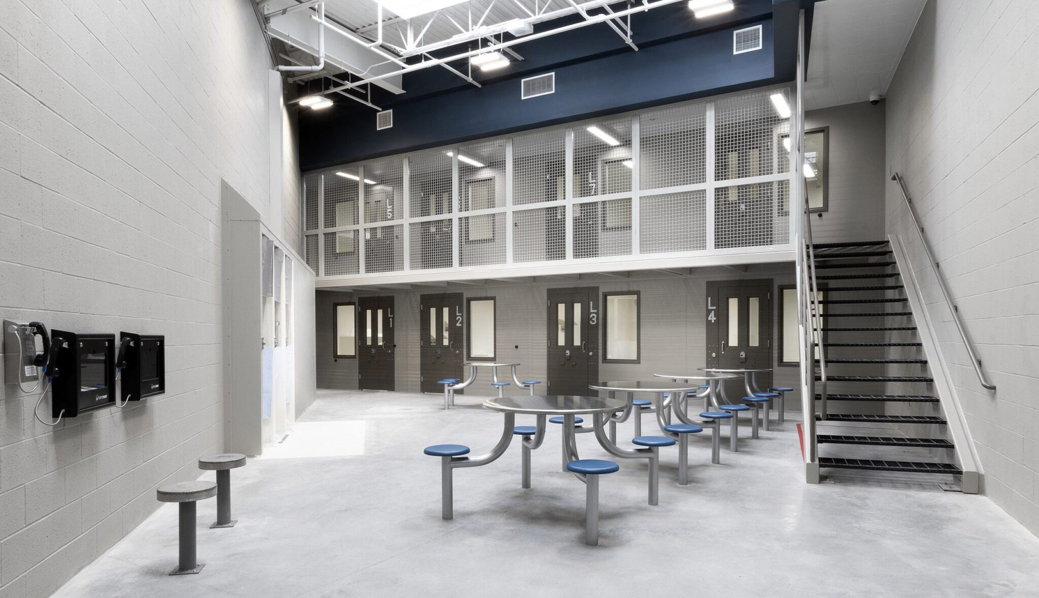 A stark, institutional space featuring metal tables, a staircase, and secure cells visible through mesh panels, with minimal decor.