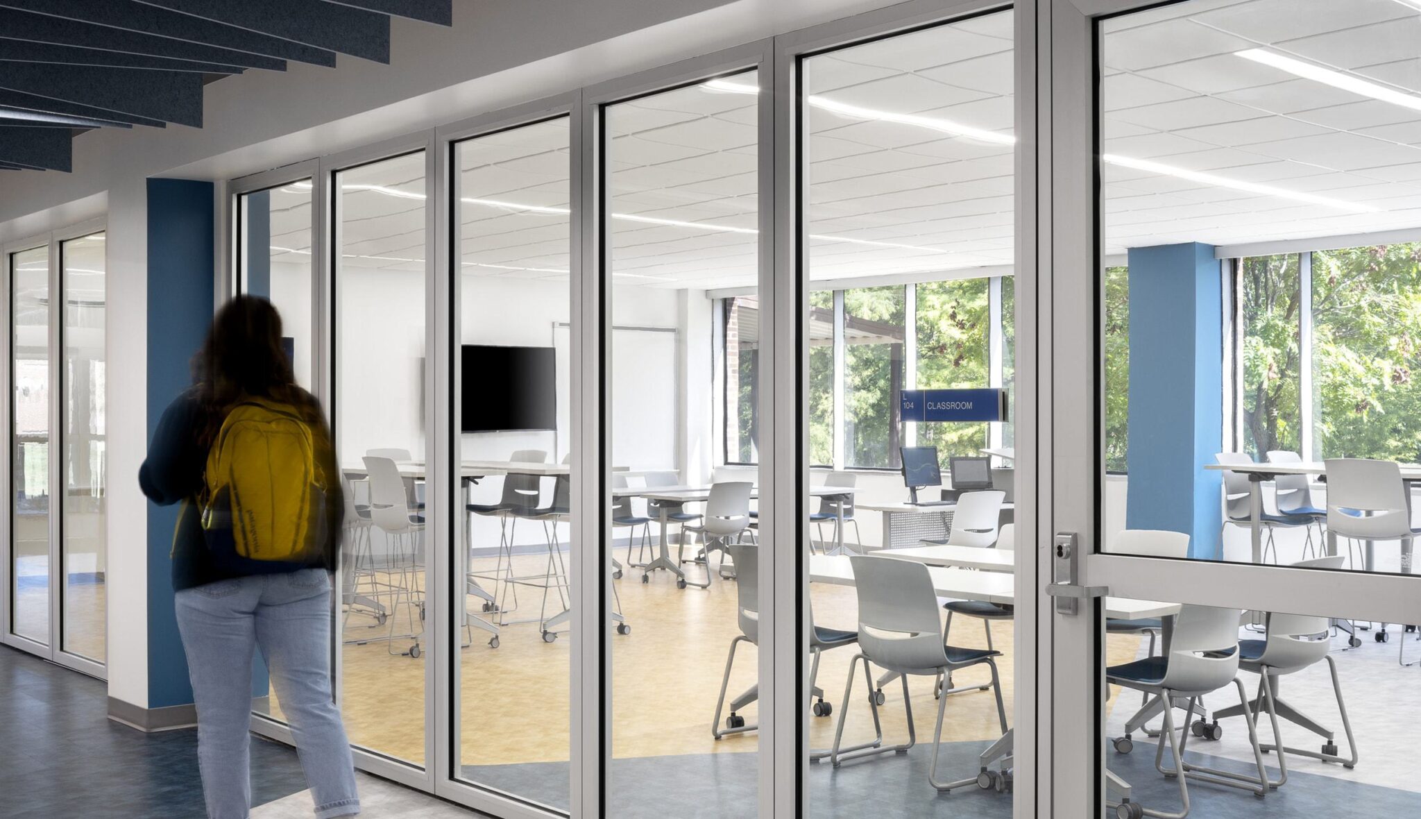 A student with a yellow backpack approaches a modern classroom with glass walls, featuring round tables and natural light.