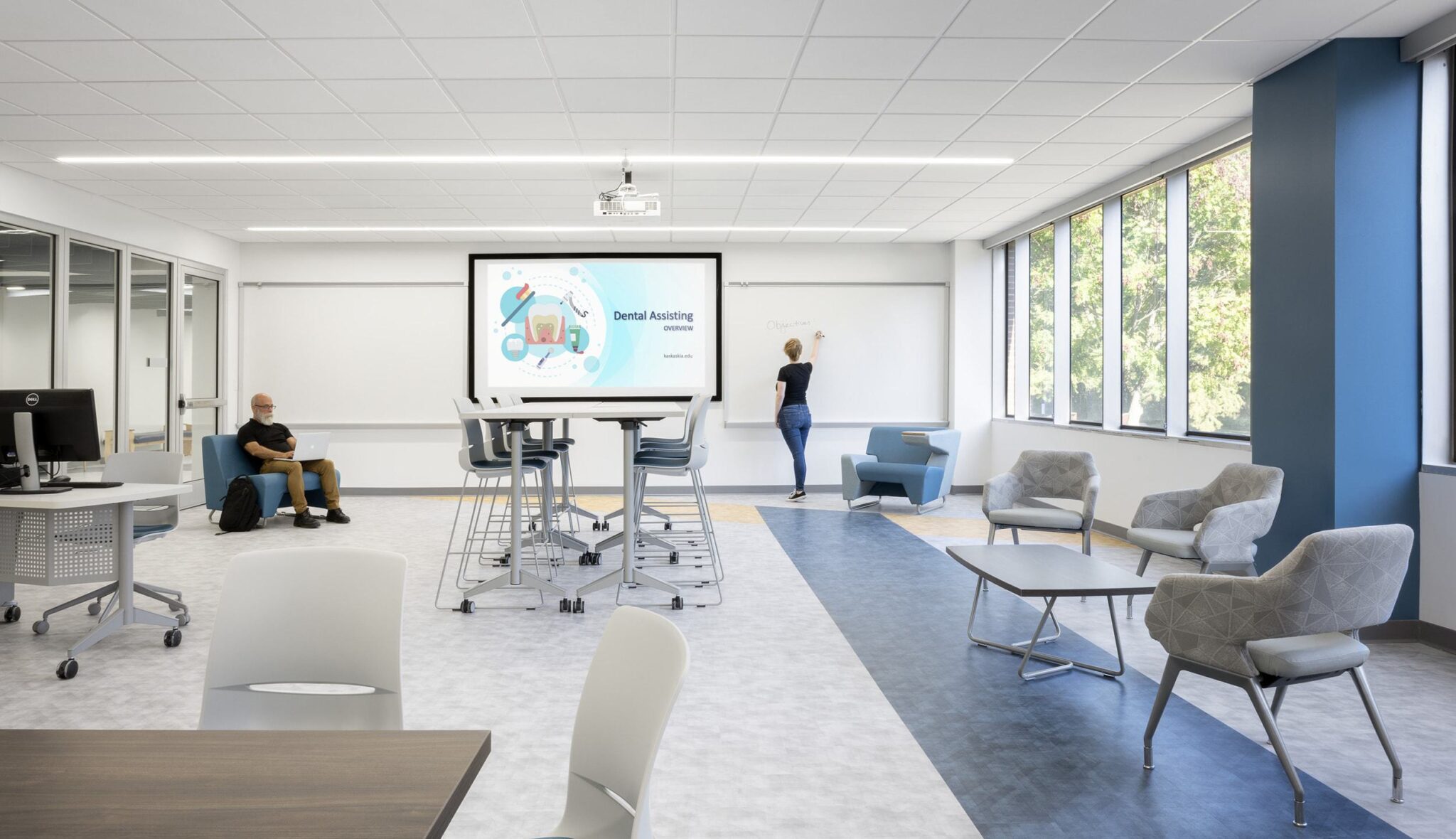 A modern learning space with collaborative seating, a large screen displaying content, and a student engaged in writing on a whiteboard.