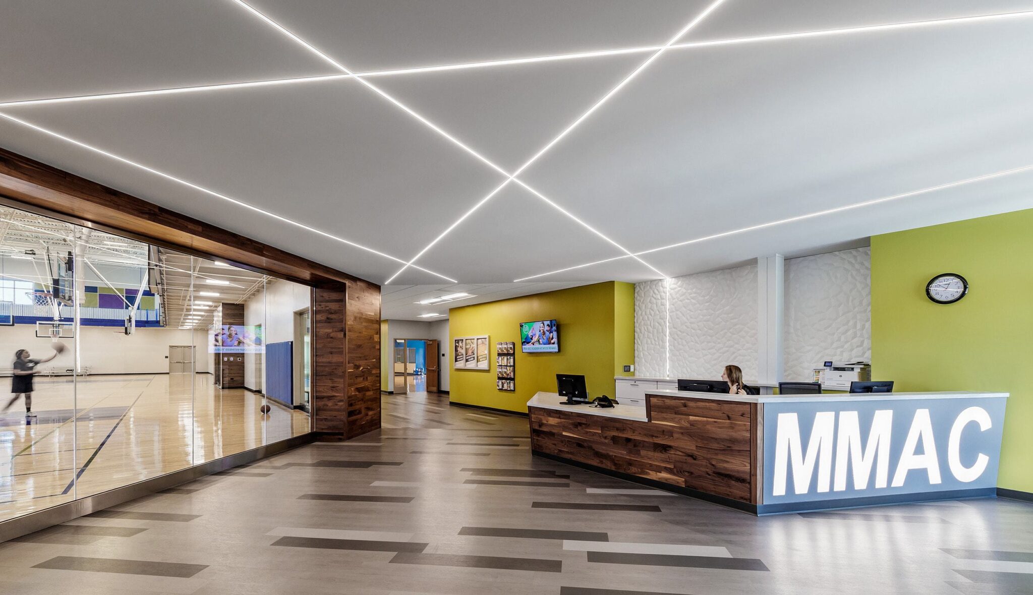 A modern recreational center lobby features a wooden reception desk, bright green walls, linear lighting, and a view of a gymnasium.