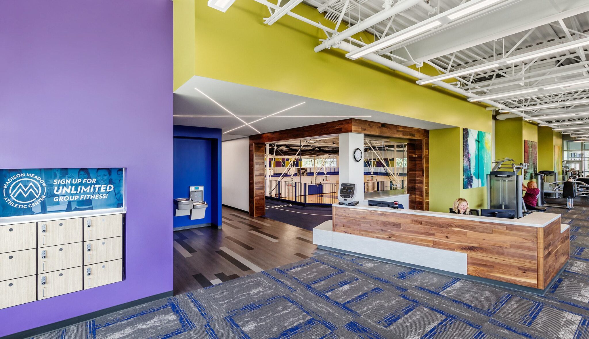 A modern reception area features colorful walls, a check-in desk, lockers, and fitness equipment in a spacious, well-lit environment.