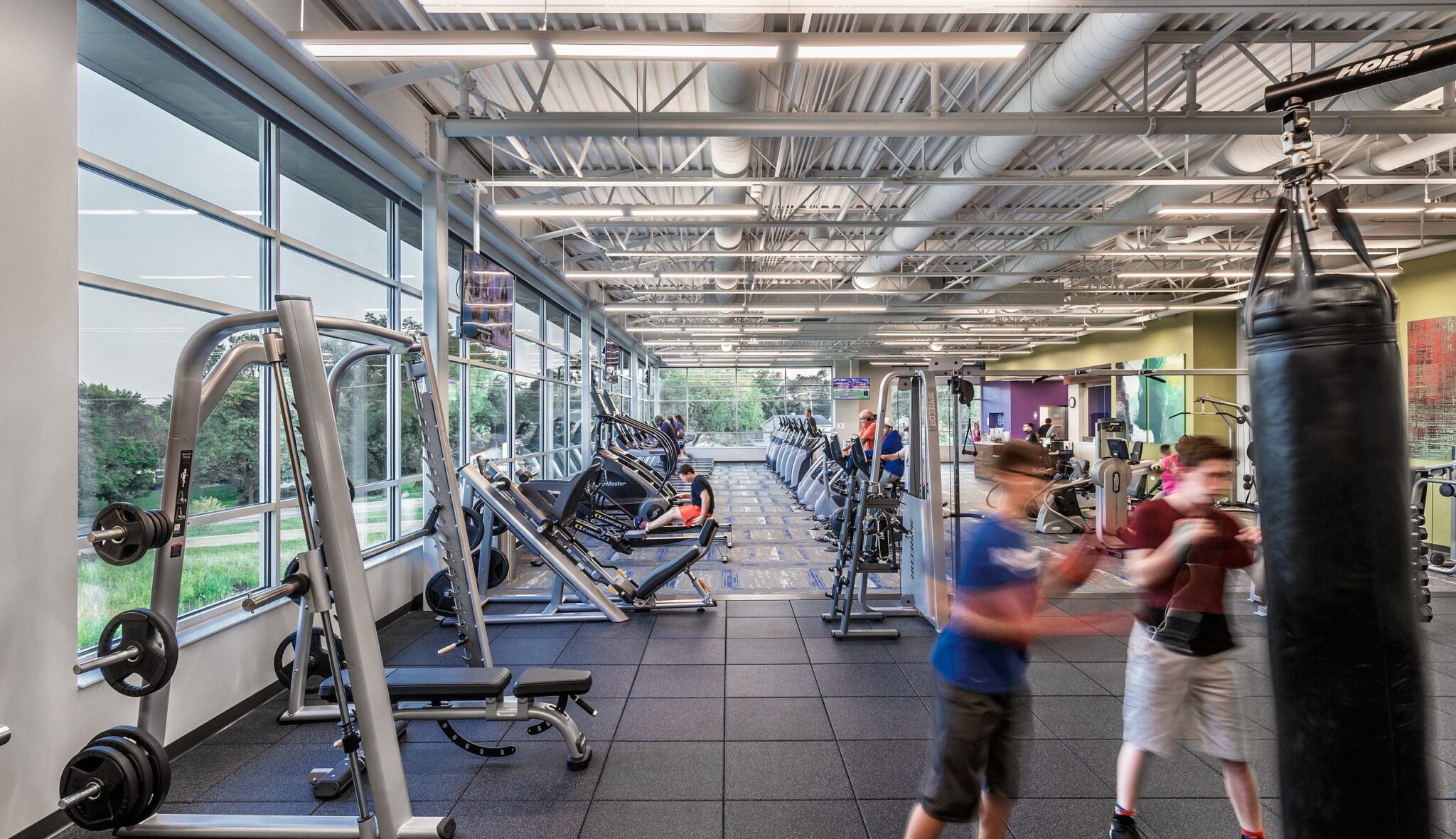 A spacious gym features various workout machines and weights, with people exercising and a punching bag in the foreground. Natural light streams in.