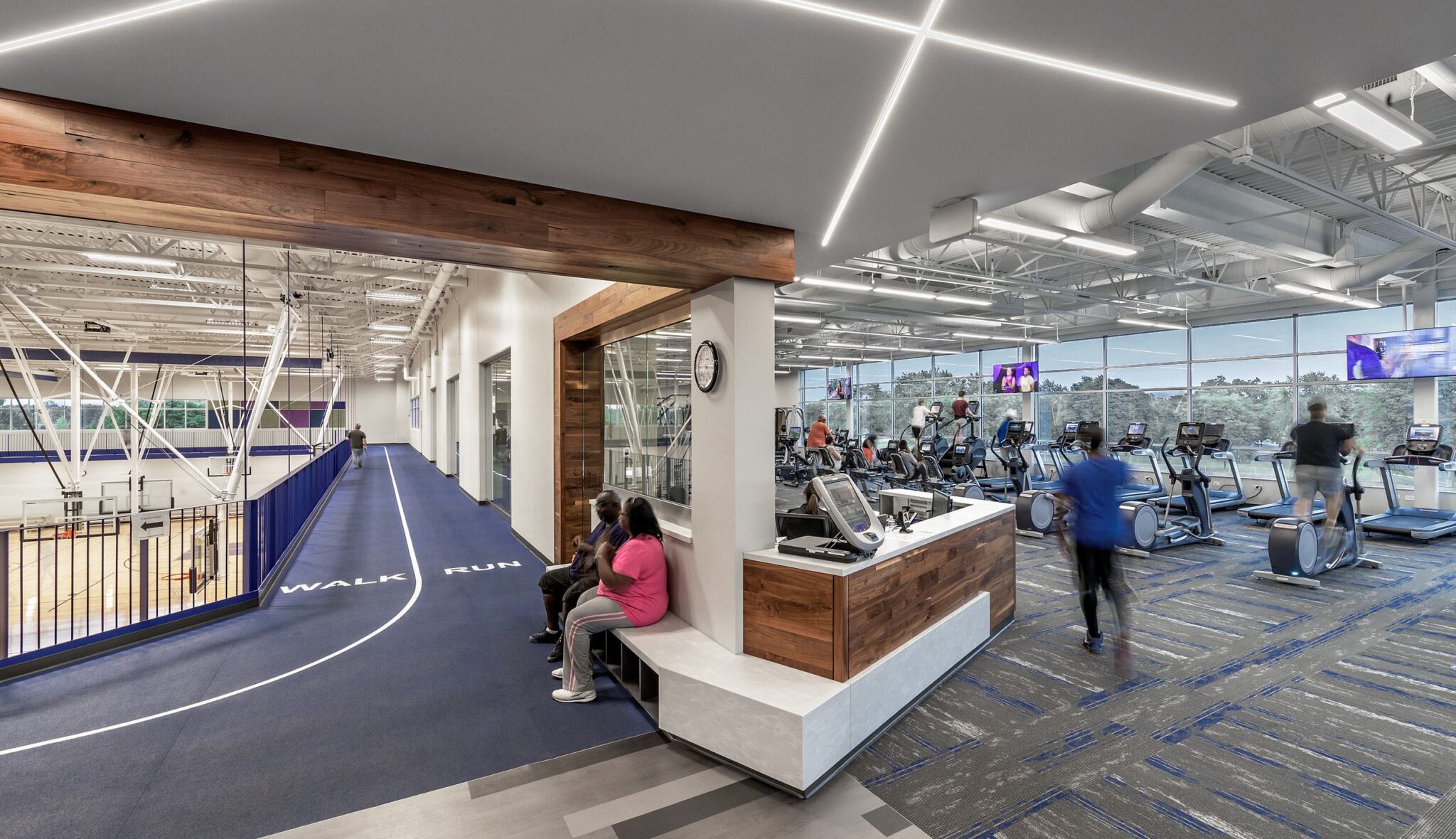 A modern gym features workout areas with treadmills, a basketball court in the background, and people engaged in various fitness activities.