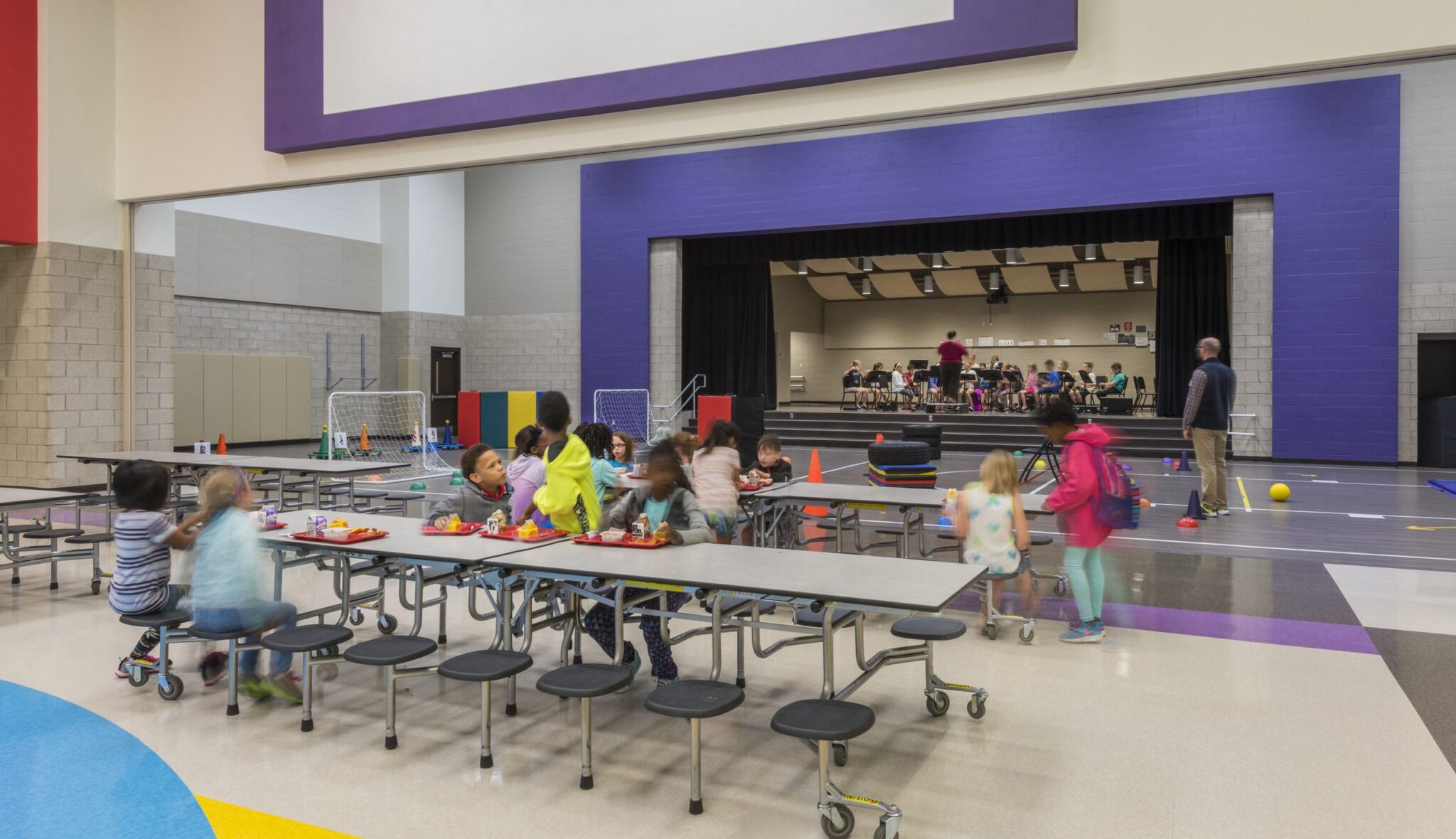 A bright school cafeteria with children sitting at tables, some eating, while others play. A stage features a group performing in the background.