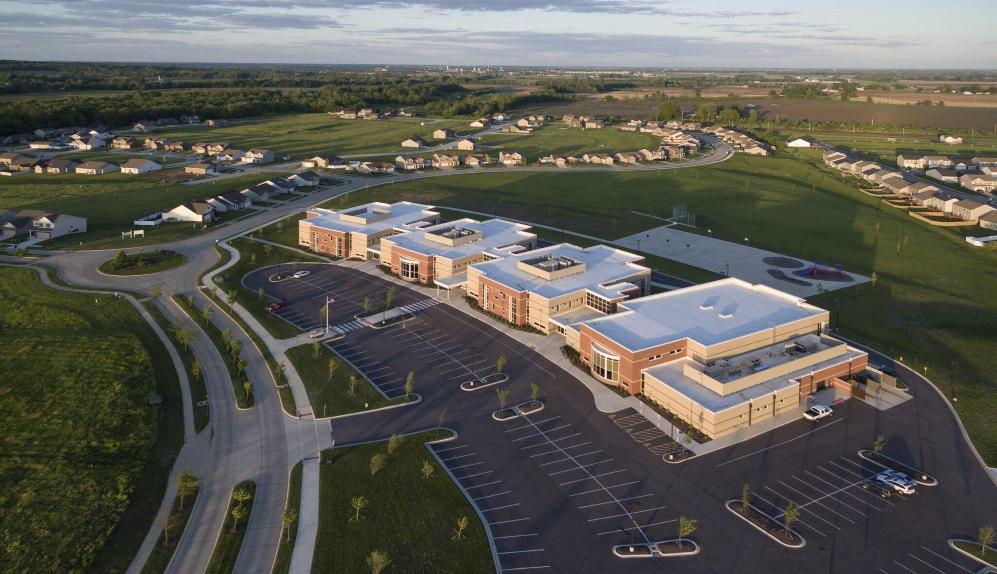 A modern building sits amidst lush green fields and residential areas, featuring ample parking and well-maintained surroundings.