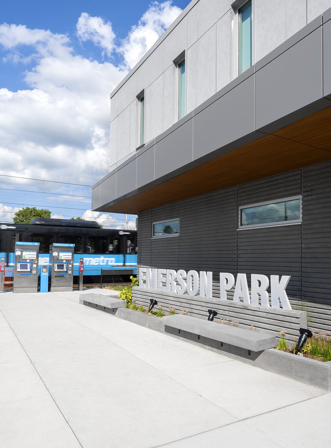 A modern transit station features "EMERSON PARK" signage, benches, and ticket machines under a bright blue sky with fluffy clouds.