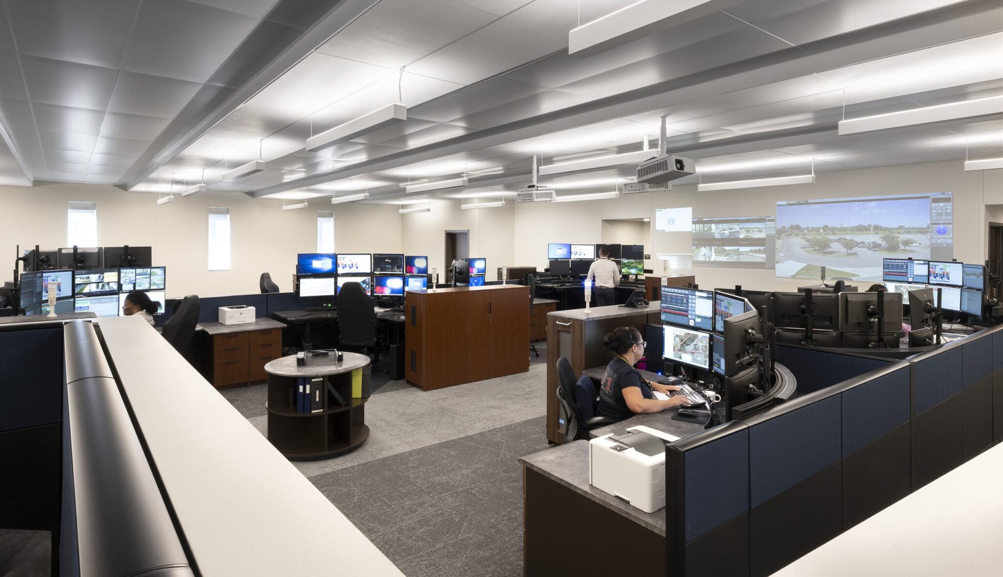 A modern control room features multiple workstations with monitors, employees engaged in tasks, and a large display projecting visuals.