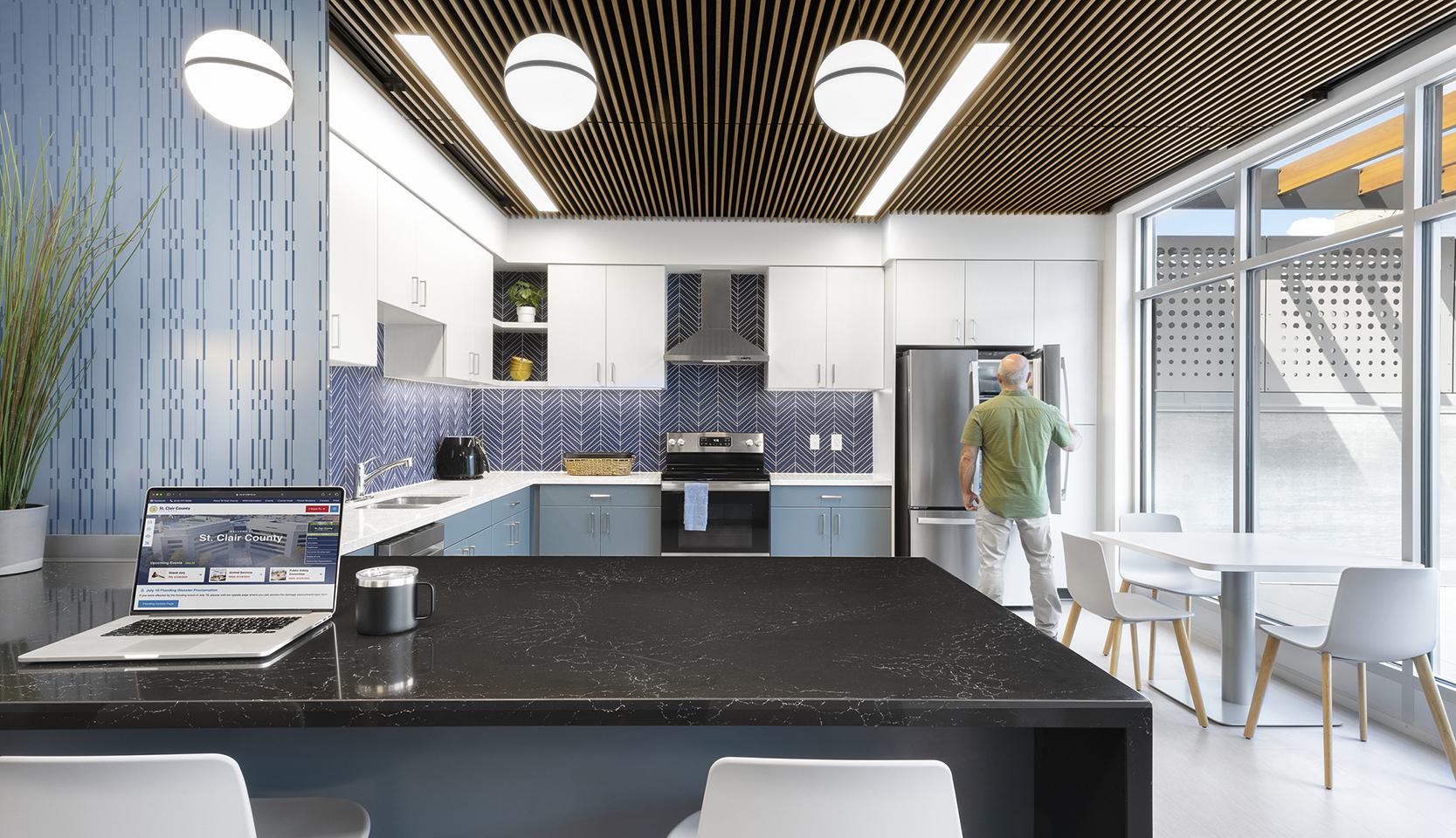 A modern kitchen features sleek cabinets, a dark countertop, and a person standing near a fridge, with bright lighting and a laptop on the island.