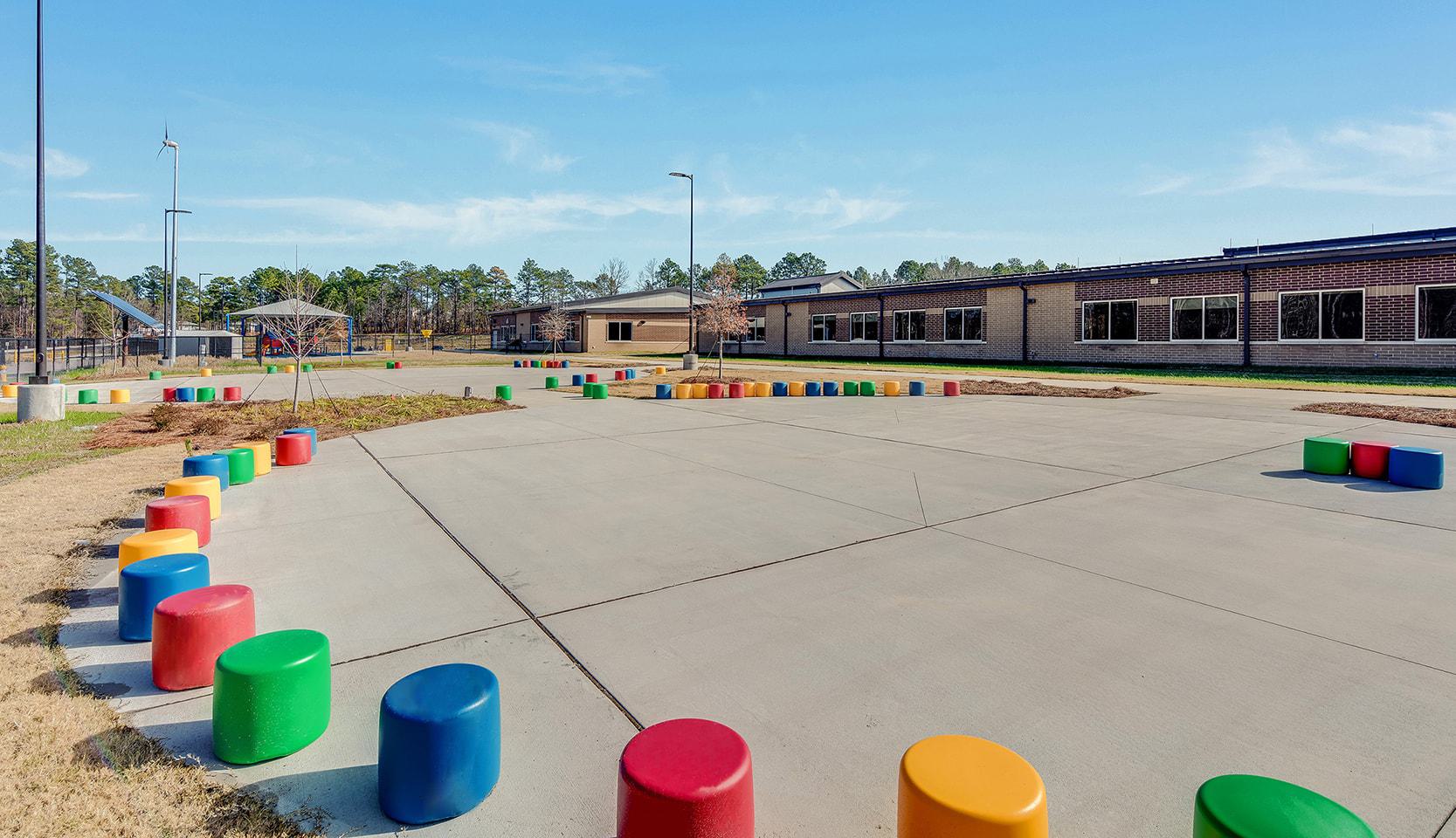 A spacious outdoor area features colorful, cylindrical seating arranged in a circle, surrounded by a grassy landscape and modern school buildings.