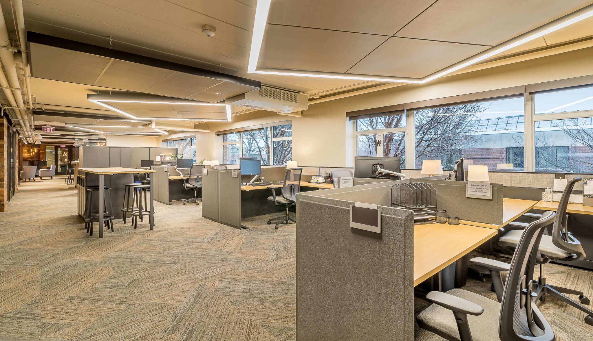 A modern, spacious office layout featuring cubicles, ergonomic chairs, and large windows providing natural light.