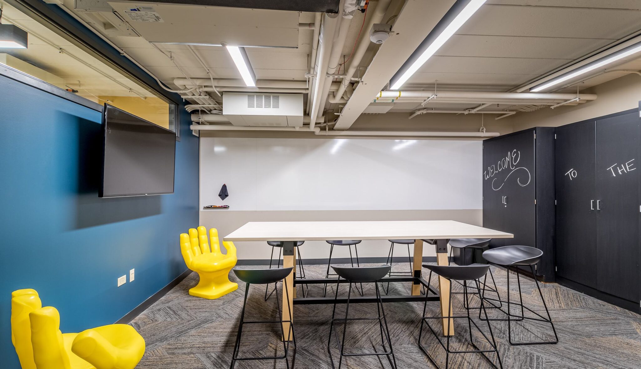 A modern meeting room featuring a large table, colorful seating, a wall-mounted TV, and a chalkboard, designed for collaborative work.