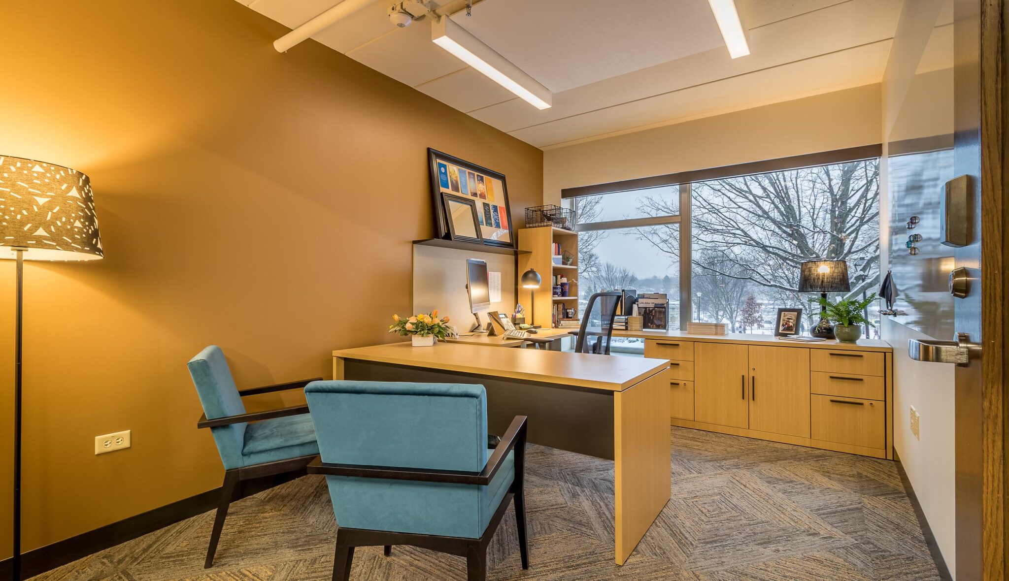 A cozy office space featuring a wooden desk, stylish blue chairs, warm lighting, and a large window overlooking a winter landscape.