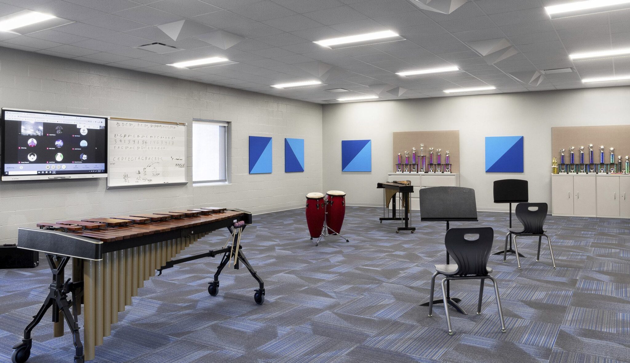 A modern music classroom features a xylophone, conga drums, chairs, and colorful wall art, with a large screen and bright lighting.