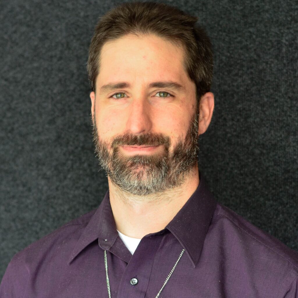 A man with a beard smiles warmly, wearing a dark purple shirt and a necklace. He stands against a textured black background.