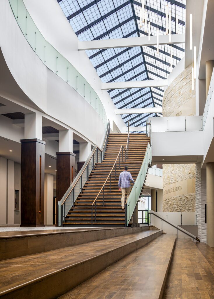 A person ascends a wooden staircase in a modern, spacious interior with large windows and elegant design elements.