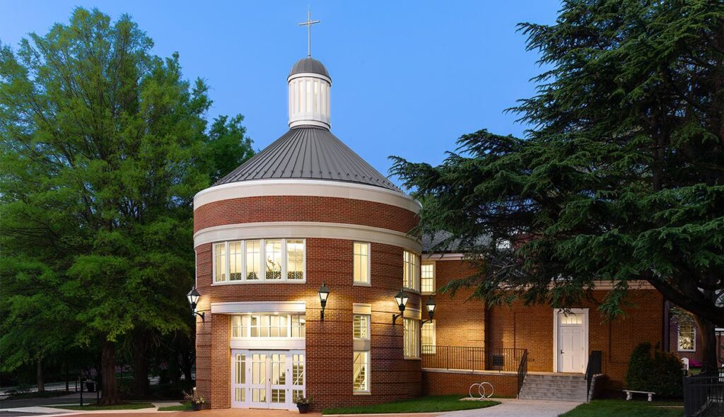 A round brick church entry with a dome and cross atop, illuminated at dusk, surrounded by trees and an inviting outdoor space.