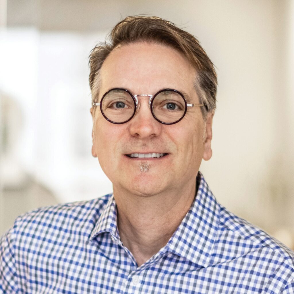 A smiling man with medium-length hair and round glasses, wearing a checkered blue and white shirt, exudes a friendly and approachable demeanor.