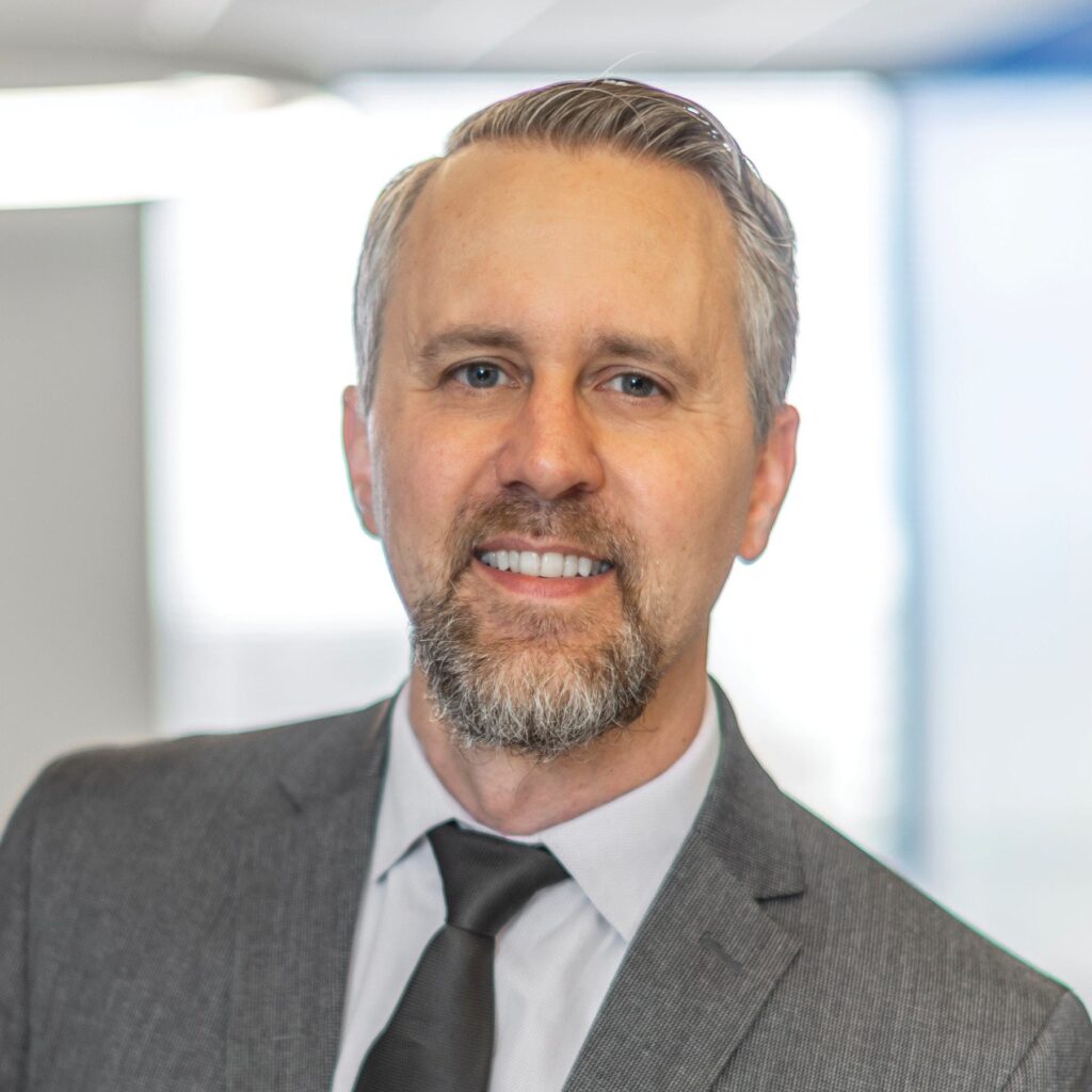 A confident professional with a light beard and neatly styled hair, dressed in a gray suit and tie, smiles warmly in a modern office setting.