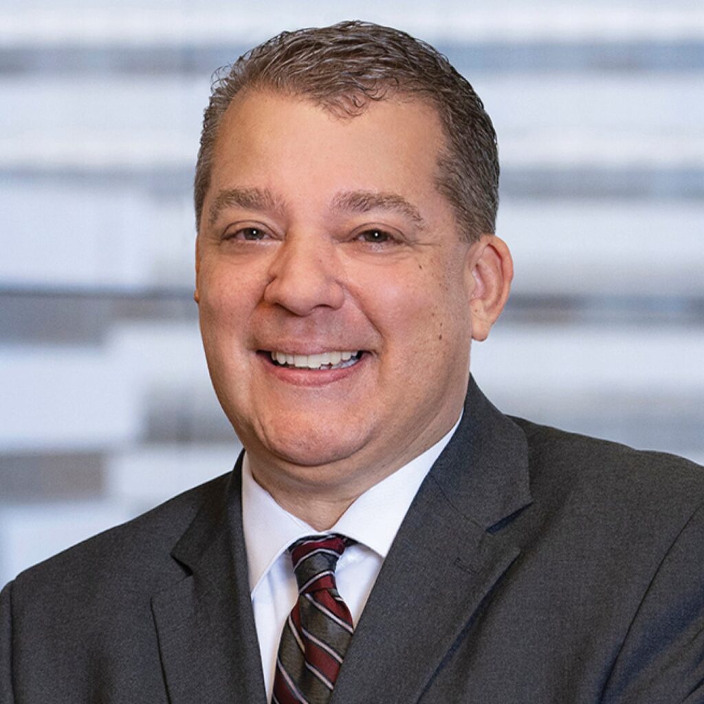 A smiling man in a suit and tie, exuding confidence and approachability, with a blurred background that suggests a professional setting.