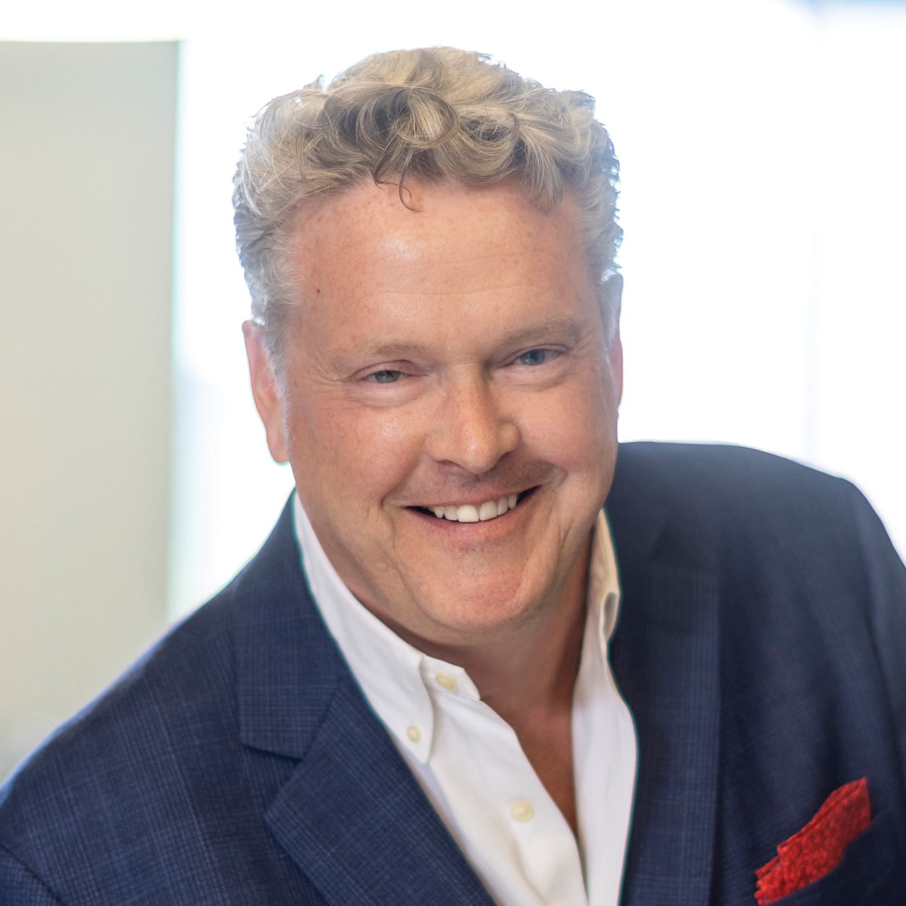 A smiling man with wavy blonde hair wears a blue blazer over a white shirt, exuding warmth and approachability.