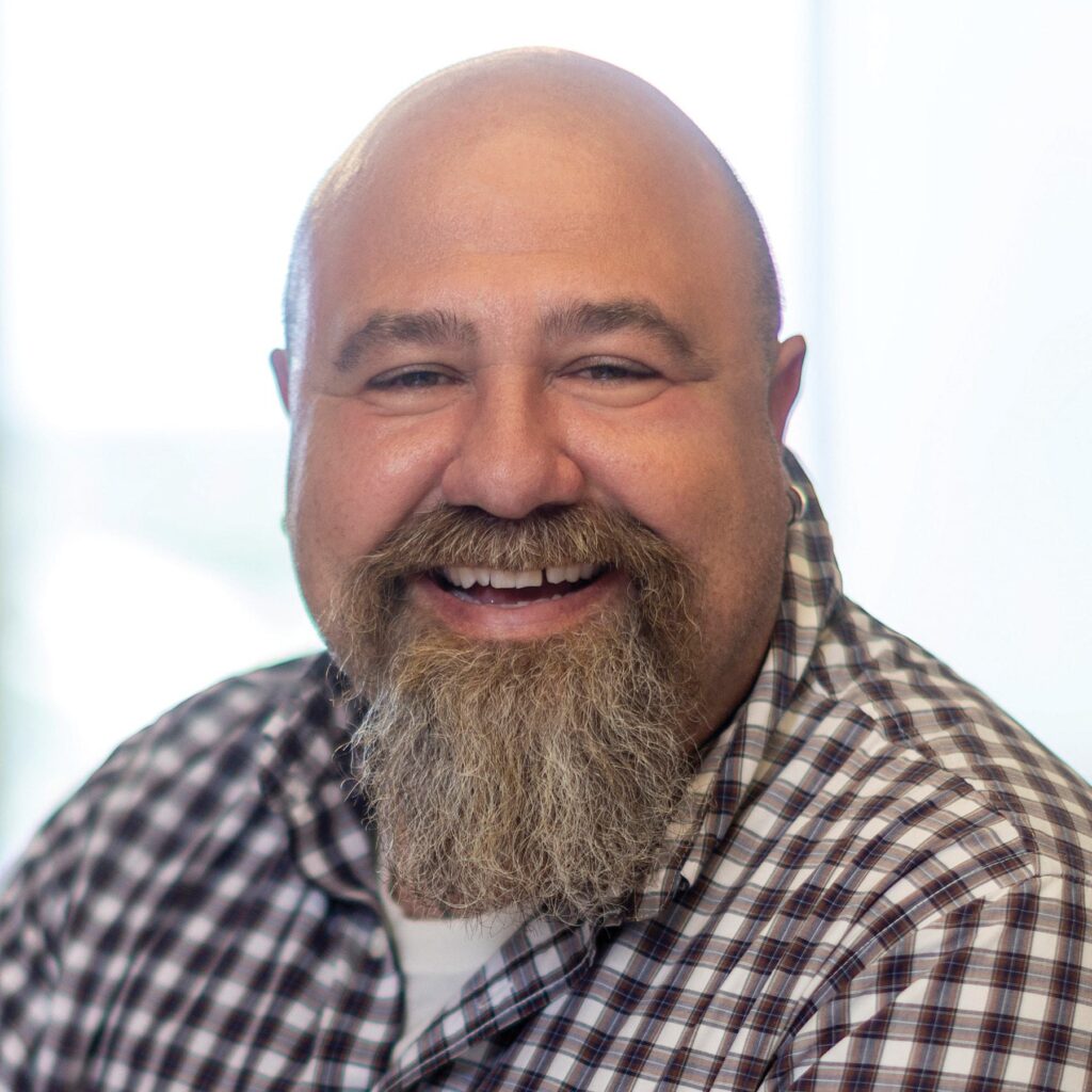 A smiling man with a bald head and a full beard, wearing a checkered shirt, exudes warmth and friendliness in a bright setting.