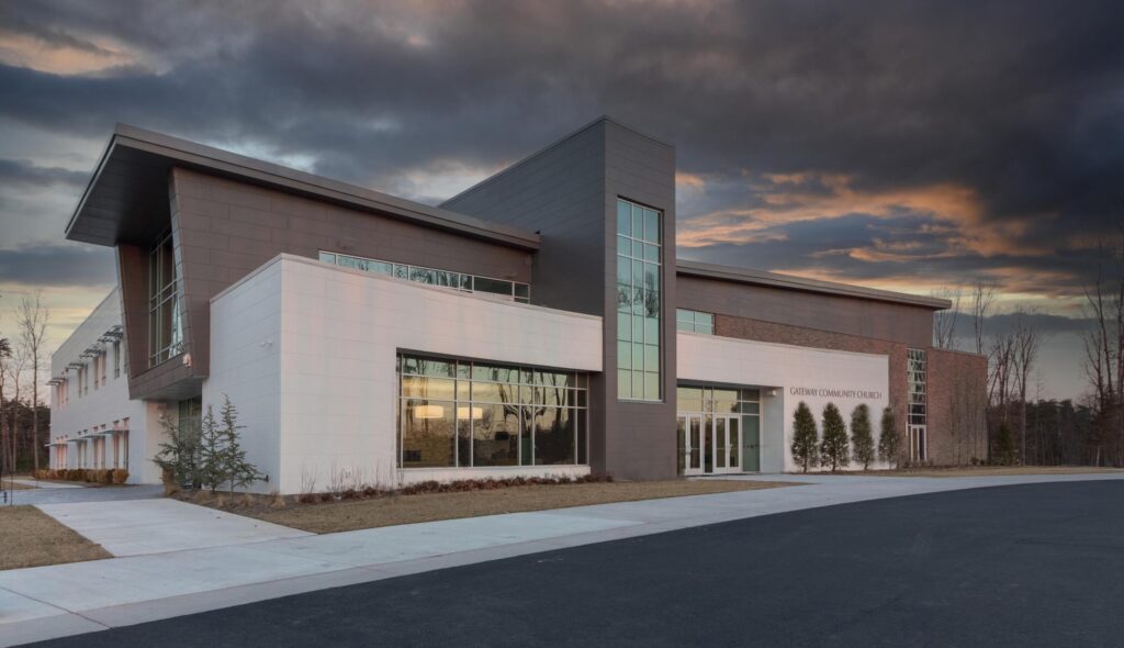 A modern church building features a blend of white and dark materials, large windows, and an open entrance, set against a dramatic sky.
