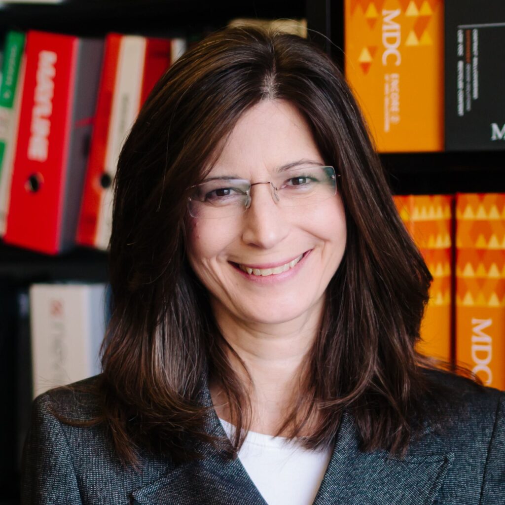 A woman with long, dark hair and glasses smiles warmly, wearing a blazer, in front of shelves filled with colorful folders.