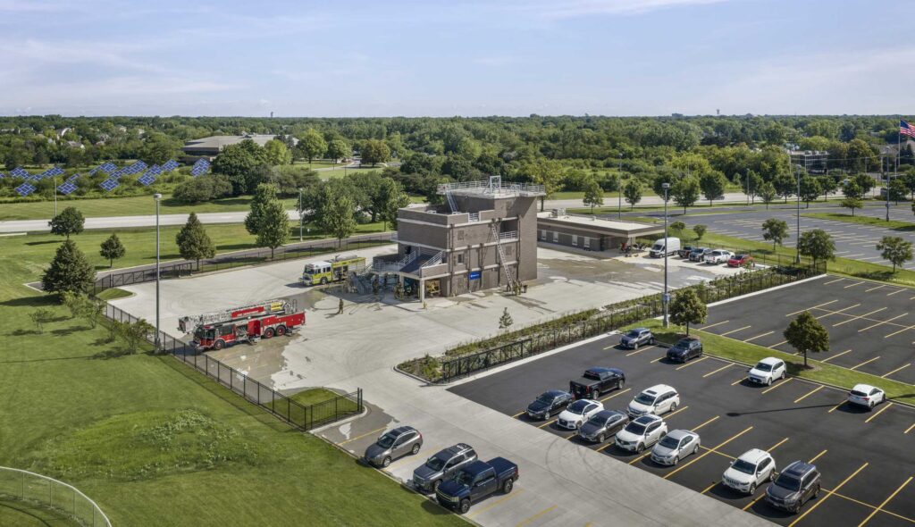 Harper College Emergency Services Training Center: Aerial view of Harper College's Emergency Services Training Center, featuring a multistory fire training tower, fire trucks, paved training grounds and adjacent parking lots, surrounded by green space and trees.