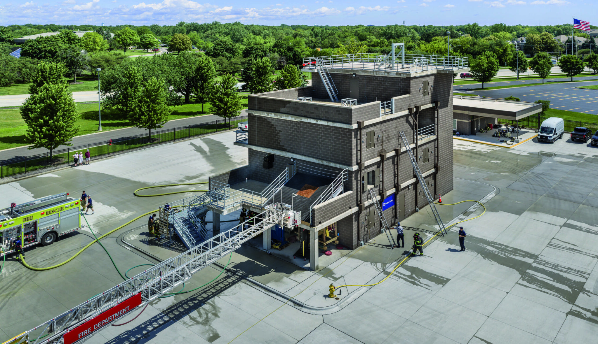 Aerial view of training tower: Elevated view of Harper College's Emergency Services Training Center showing the multistory fire training tower, exterior stairways, rooftop platforms and an extended fire truck aerial ladder during training exercises.
