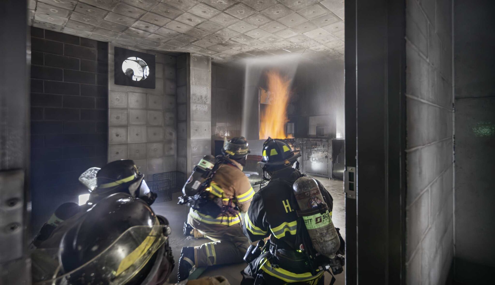 Interior fire training: Firefighters wearing full protective gear and breathing apparatus practice fire suppression techniques inside a smoke-filled training room at Harper College's Emergency Services Training Center, with flames rising from a controlled fire prop.