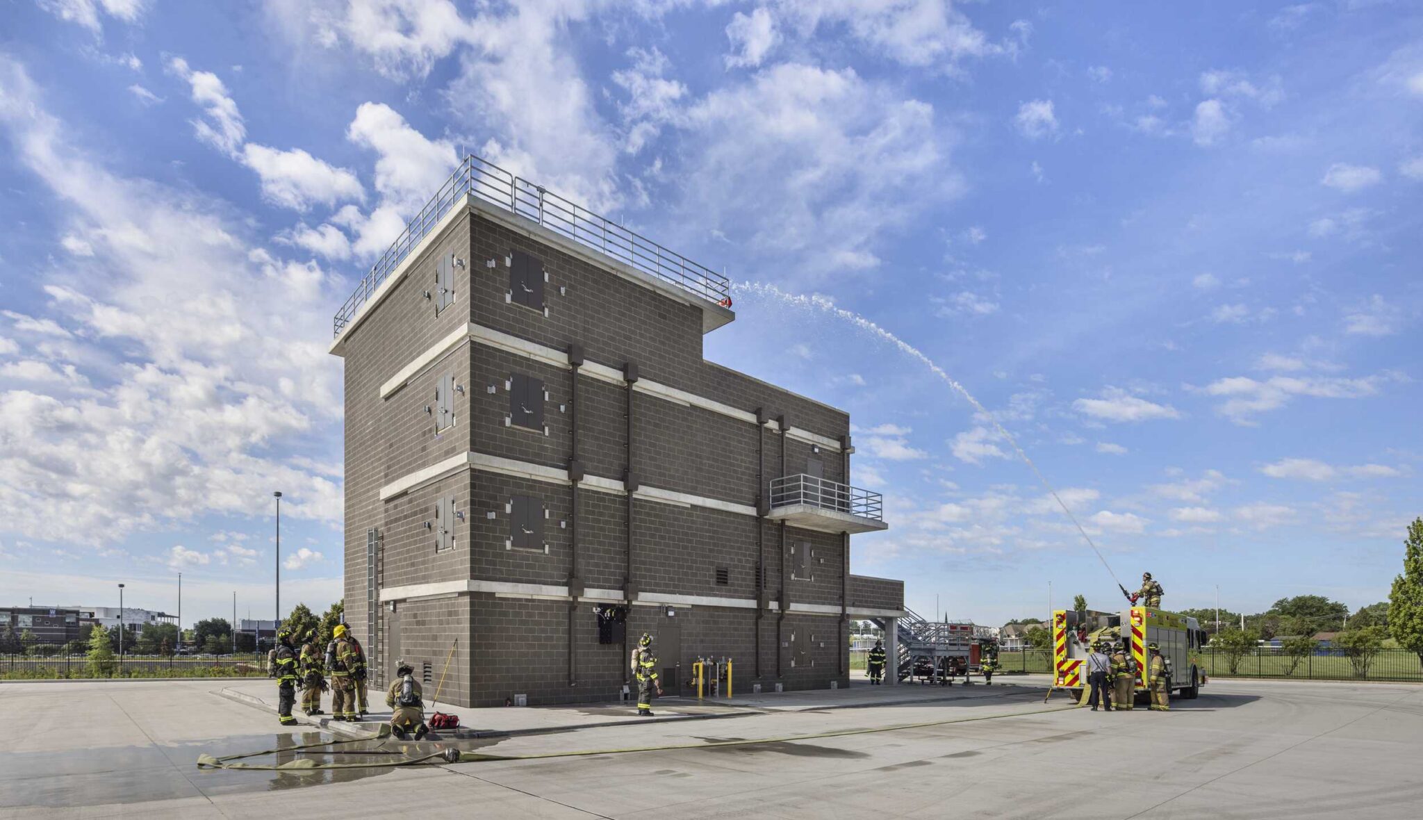 Fire training tower exterior: Firefighters conduct training exercises around Harper College's multistory fire training tower, featuring concrete-block construction, exterior stairways, elevated platforms and a fire truck with hoses deployed.