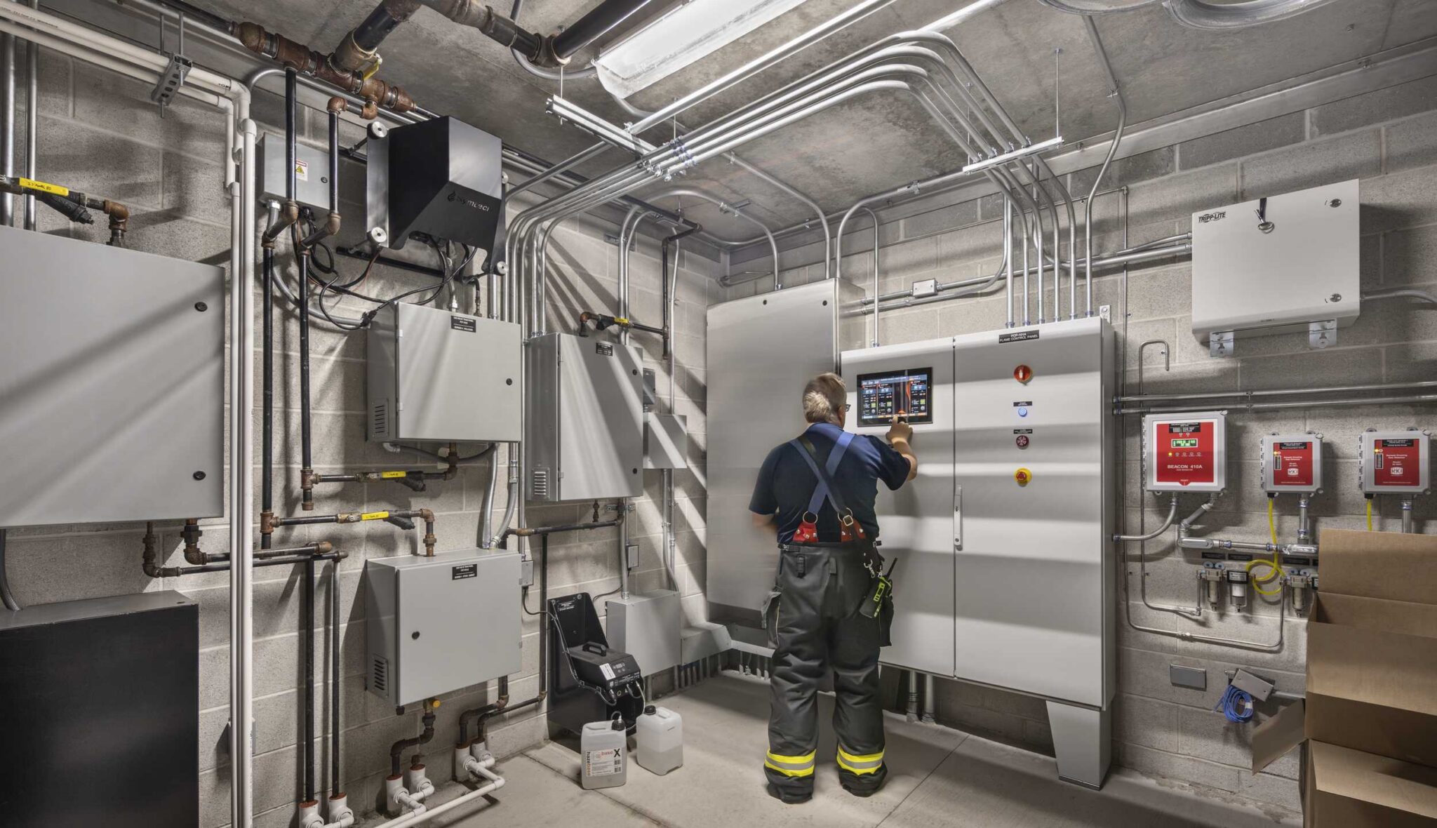 Fire training control room: A staff member operates a digital control panel inside Harper College's Emergency Services Training Center, surrounded by electrical cabinets, exposed piping and monitoring equipment.
