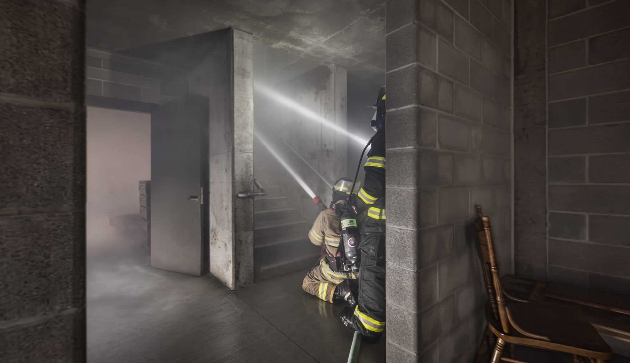 Firefighter stairwell training: Two firefighters wearing protective gear and breathing apparatus practice hose operations in a smoke-filled stairwell inside Harper College's fire training tower.