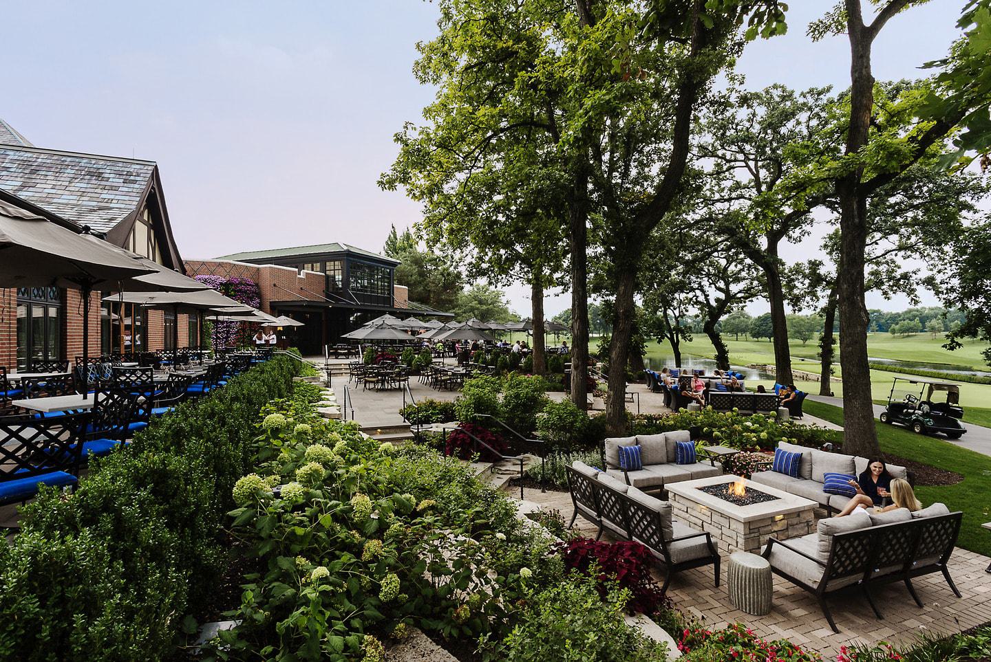 A serene outdoor dining area features cozy seating around a fire pit, surrounded by lush greenery and a scenic view of the golf course.