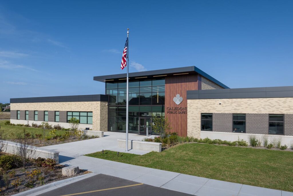 A modern public library features a mix of stone and glass architecture, surrounded by landscaped greenery and an American flag flying prominently.