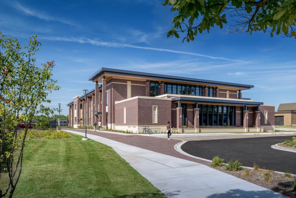 A modern, brick building with large windows surrounded by greenery and a paved walkway, under a clear blue sky.
