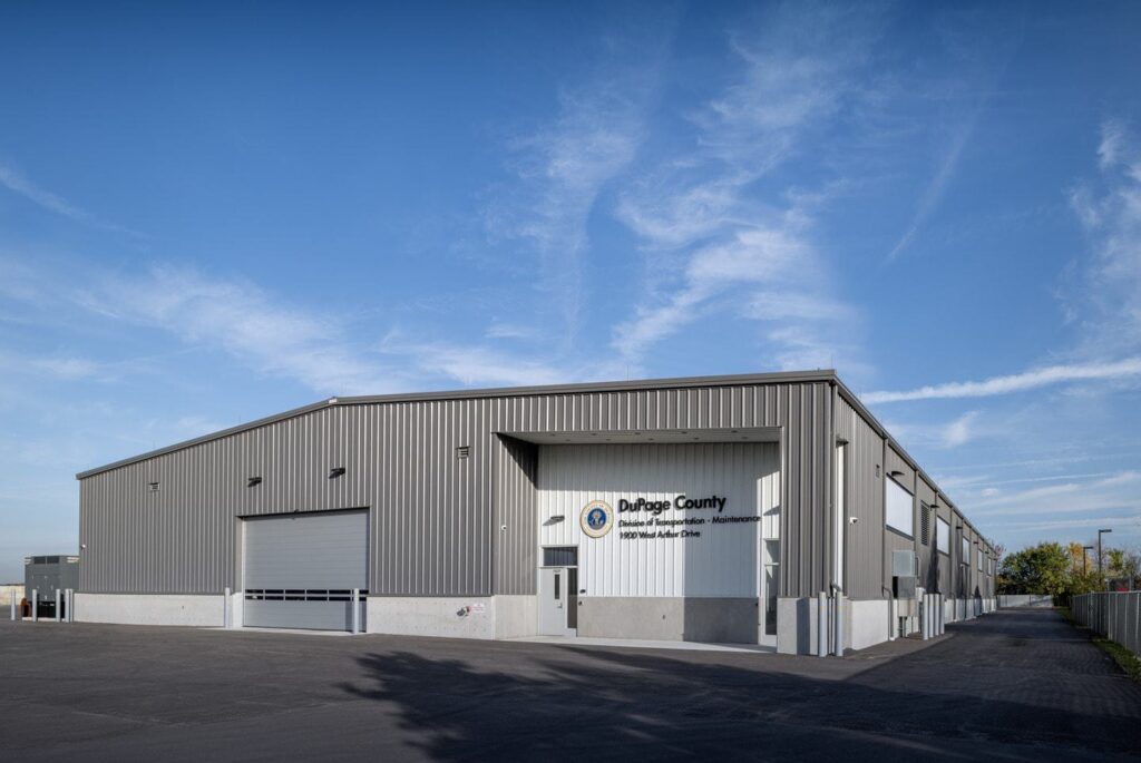 A large, modern industrial building features a gray metal exterior, a spacious loading area, and clear blue skies above.