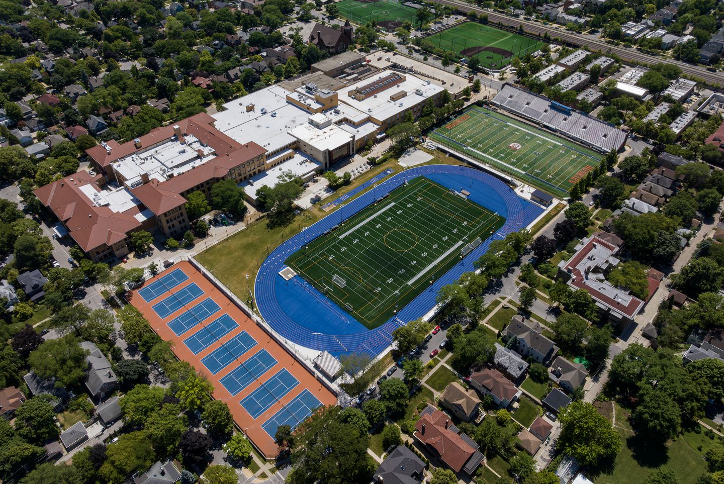 Aerial view of a school complex featuring a football field, tennis courts, and nearby athletic fields, surrounded by residential areas.