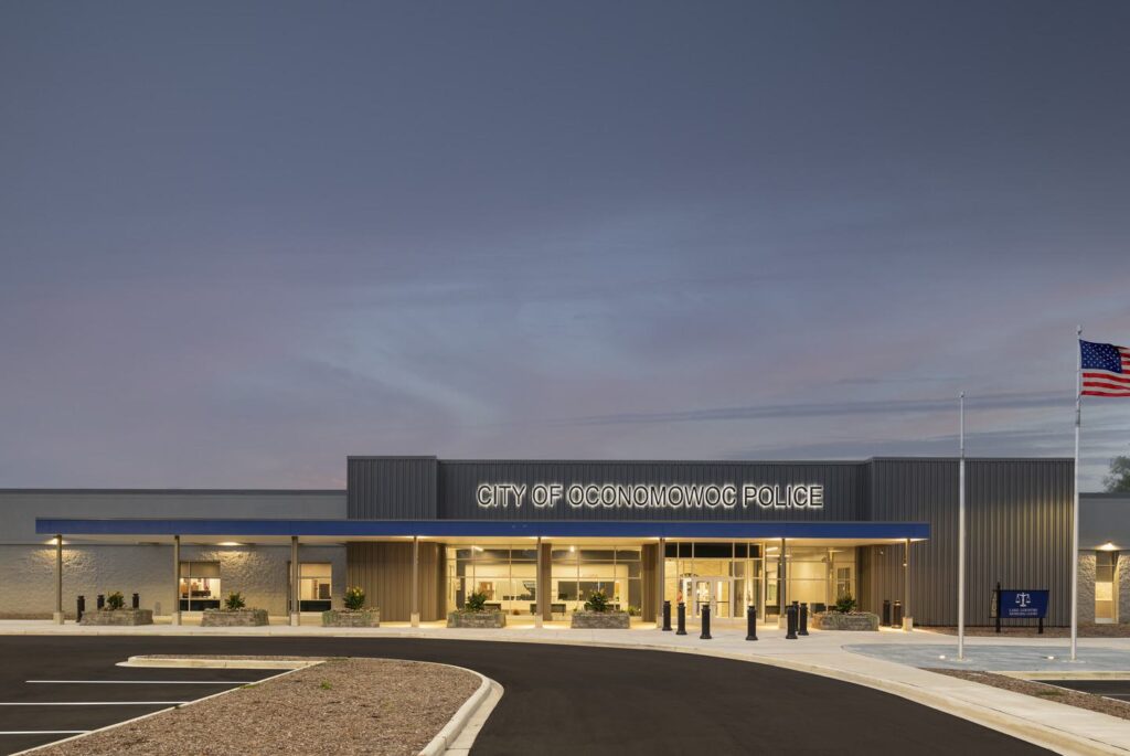 A modern police station features a sleek design with large glass entrances, landscaping, and an American flag, set against a twilight sky.