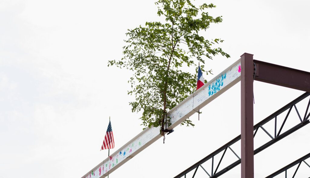 Small tree sits on top of a signed steel beam at the highest point of a building's steel frame, marking the topping out point in construction. Small sapling tree is sandwiched between an American flag and a Texas state flag. Photo courtesy of Rogers-O'Brien Construction.